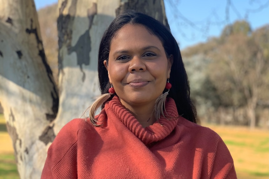 Lynda is smiling at the camera with her arms crossed and is wearing an orange wool jumper