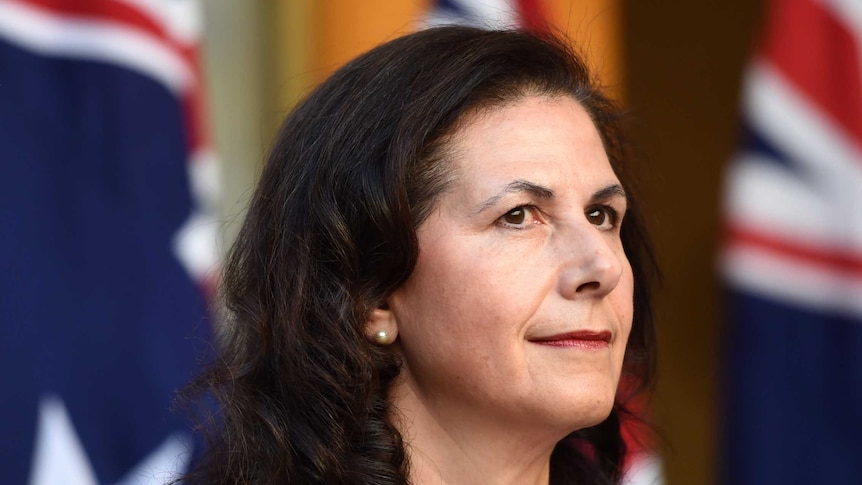 Senator Concetta Fierravanti-Wells is wearing a pearl necklace and earrings and standing in front of several Australian flags.