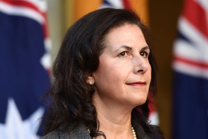 Senator Concetta Fierravanti-Wells is wearing a pearl necklace and earrings and standing in front of several Australian flags.