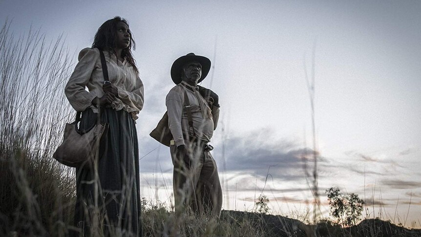 The two actors stand, in tattered period garb, on a scrub covered hill in front of a bright sunset.