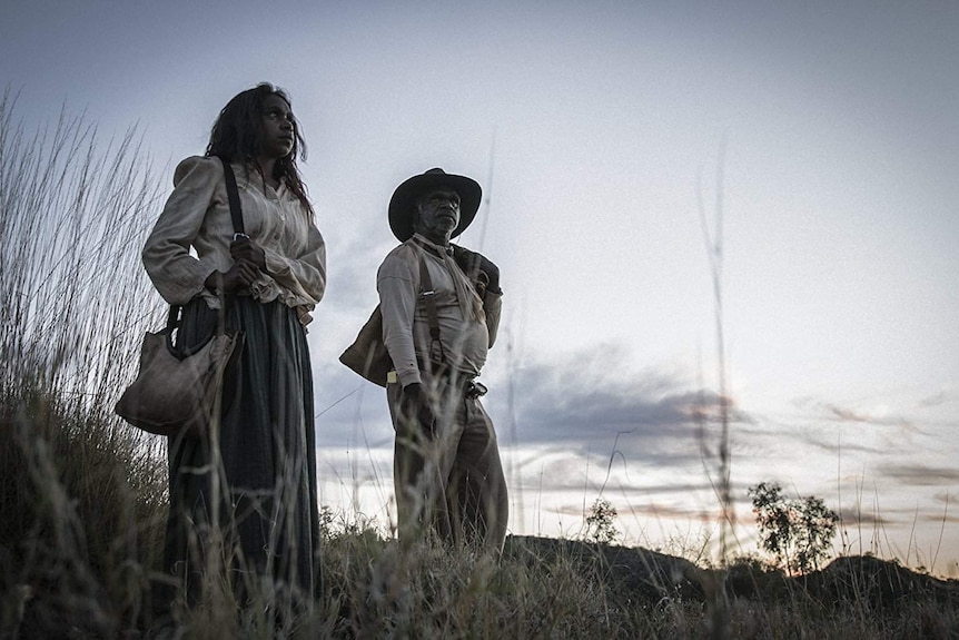 The two actors stand, in tattered period garb, on a scrub covered hill in front of a bright sunset.