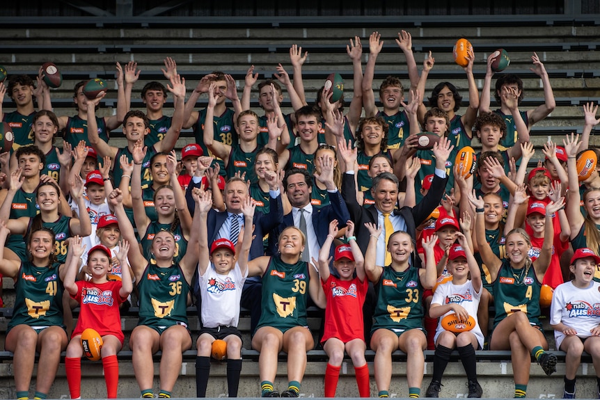 Два политика и глава АФТ окружены детьми и молодыми людьми, одетыми в футбольную форму Аускика или Тасмании.