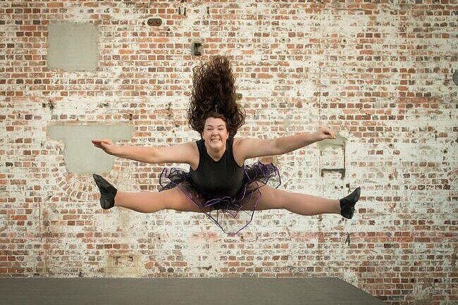 A ballerina wearing black suit does the splits in the air facing the camera with a brick wall behind her
