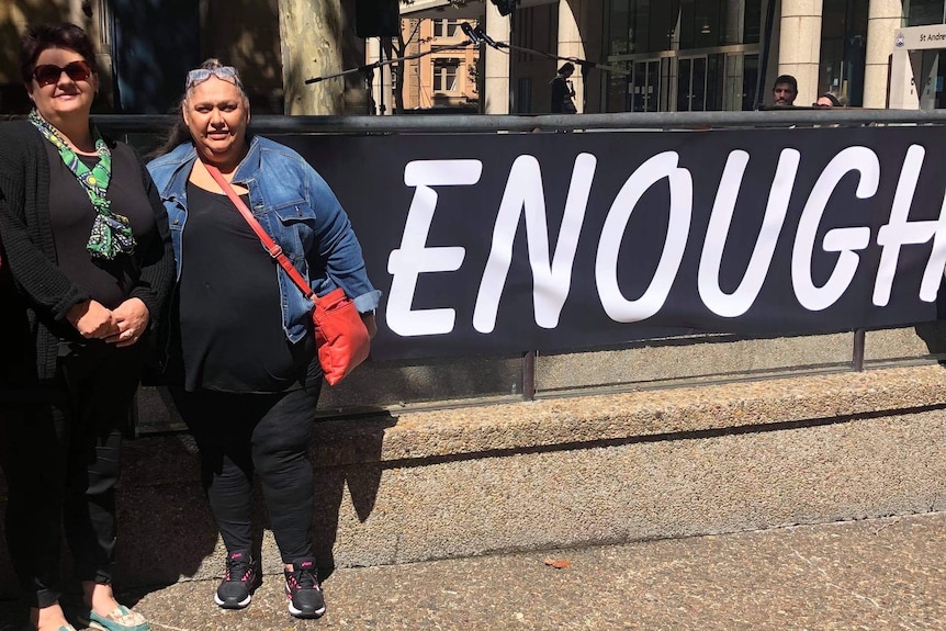 Two women stand in front of a sign that says "enough".