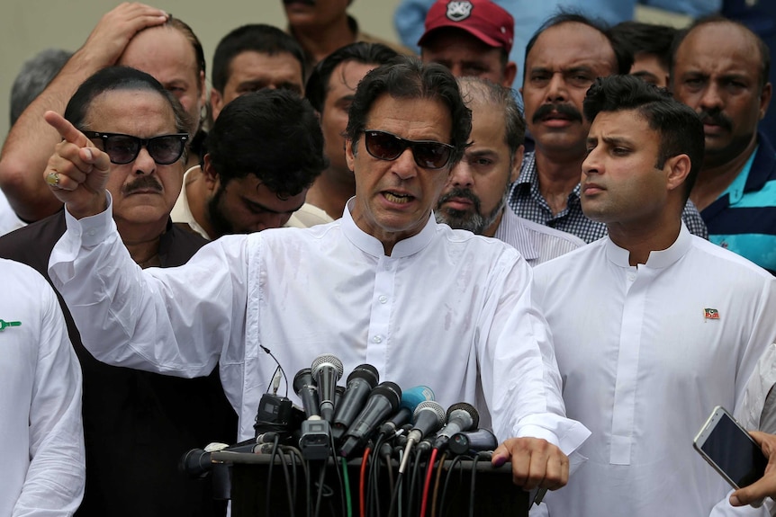 Imran Khan, wearing sunglasses, gestures as he speaks at a podium, surrounding by other men.