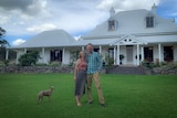 Man, woman and dog standing in front of country homestead.