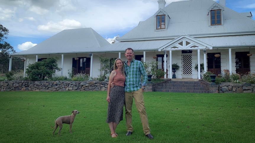 Man, woman and dog standing in front of country homestead.