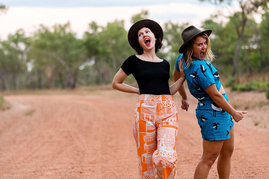 Two young women in bright clothing pose on a dirt road with scrub behind them enjoy working and living in regional Australia.