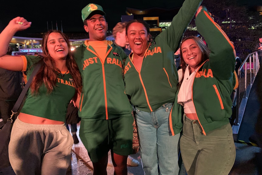 four young people cheering and raising their arms at a fan site watching a football game