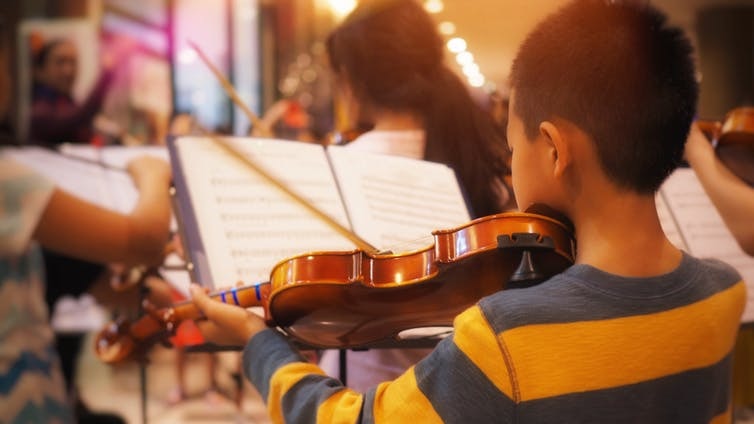 A child from behind playing violin with other children playing violin out of focus in the background.