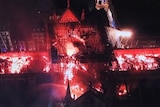An aerial shot shows bright flames burning through the roof frame of a large cathedral while firefighters spray water
