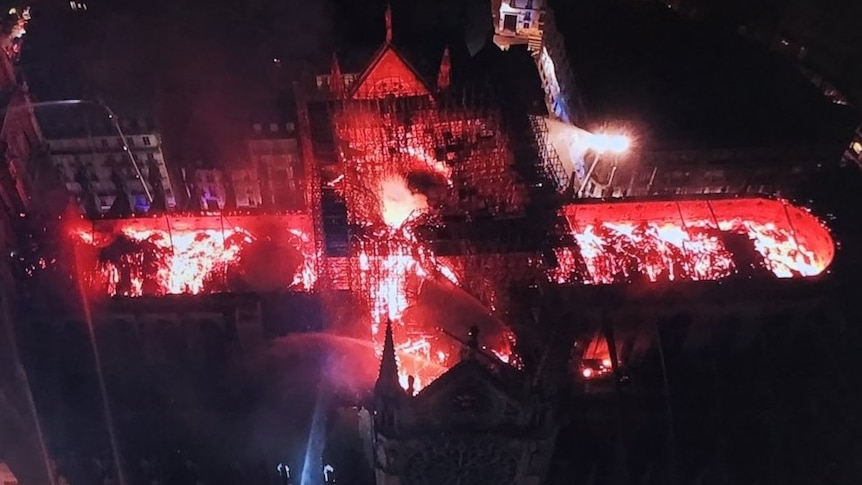 An aerial shot shows bright flames burning through the roof frame of a large cathedral while firefighters spray water