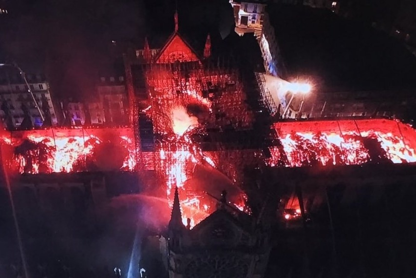 An aerial shot shows bright flames burning through the roof frame of a large cathedral while firefighters spray water