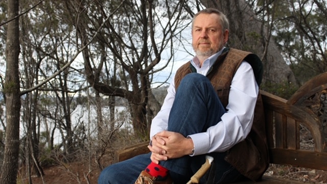 A man sits on a park bench with bushland in the background