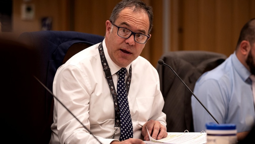 A man in a white shirt sits at a desk and speaks