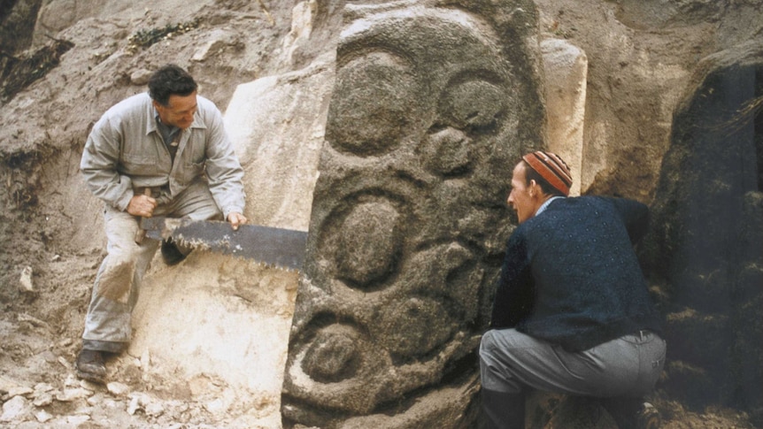 Two men stand by a rock with circular carvings on it. They are using a saw to separate it from a cliff-like structure