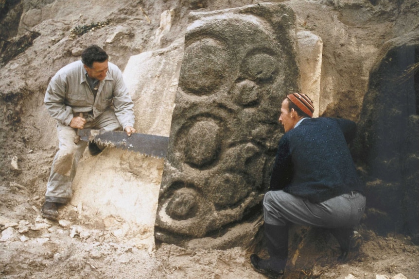 Two men stand by a rock with circular carvings on it. They are using a saw to separate it from a cliff-like structure