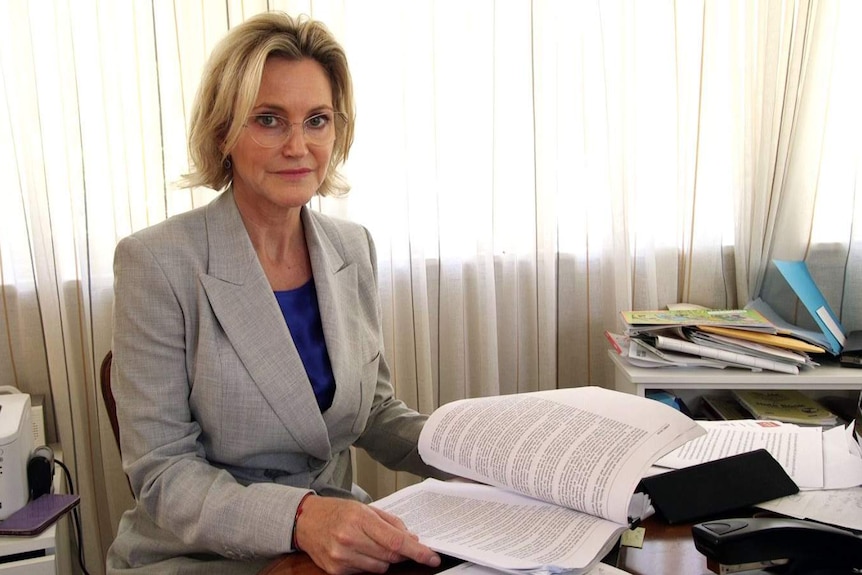 Ms Parke sits at a desk, holding a sheaf of documents.