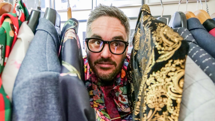 A man wearing glasses pokes his head between a rack of colourful jackets.