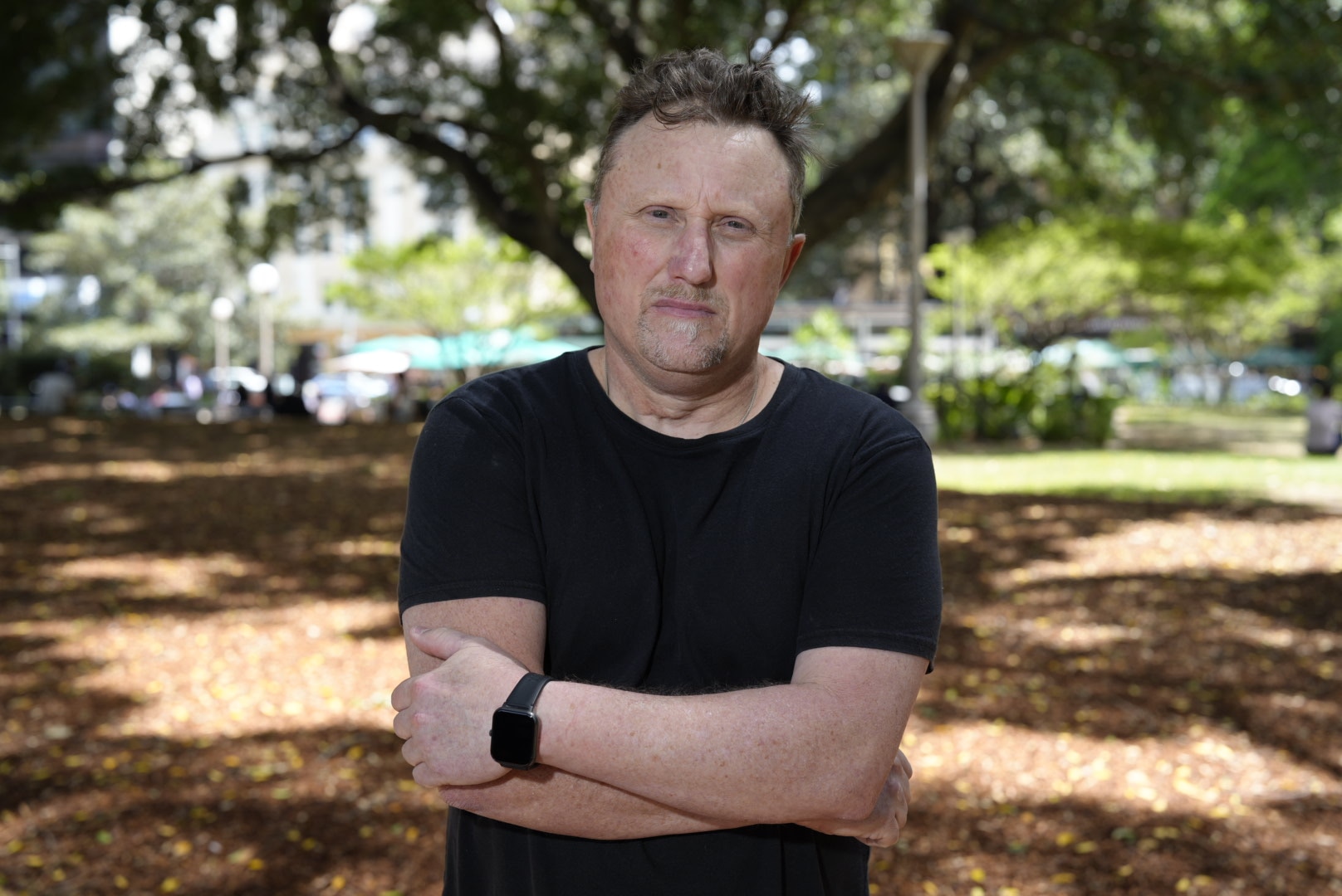Adam Watts, arms crossed, wearing a black shirt, standing in a park.
