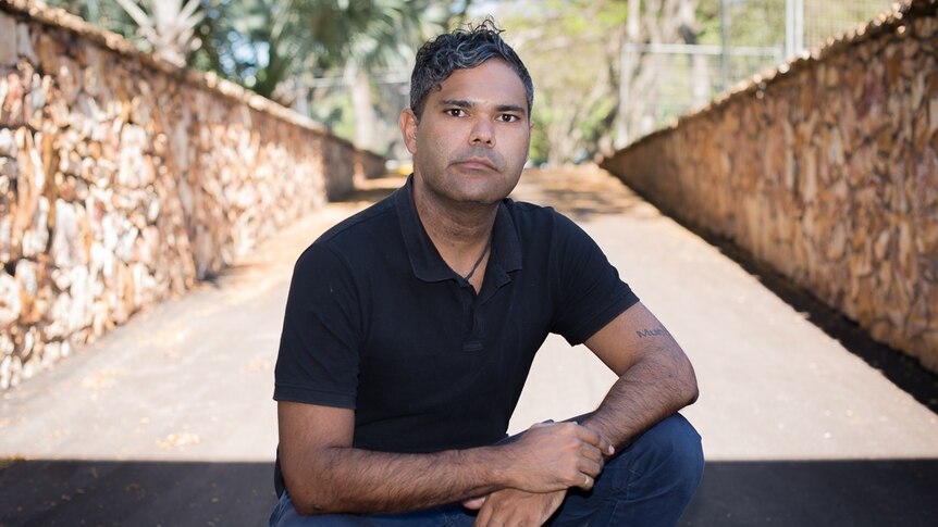 An Indigenous man kneels seriously before the camera with stone walls either side of him.