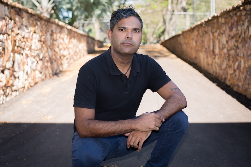 An Indigenous man kneels seriously before the camera with stone walls either side of him.