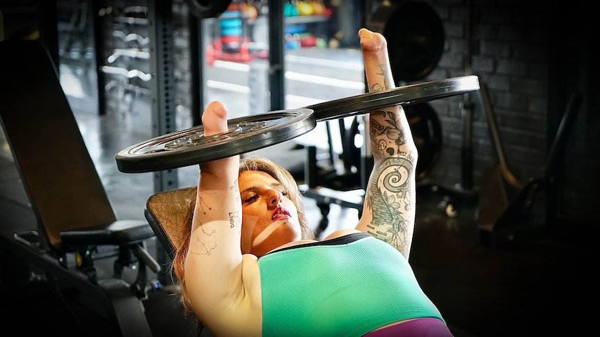 A woman in a green singlet lefts two round weights above her head at the gym. She has no hands and lies on a bench