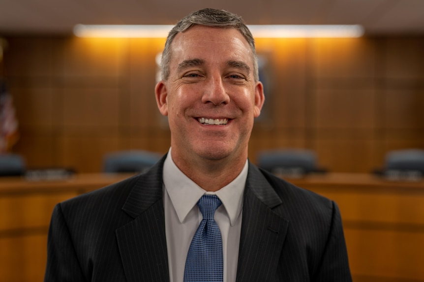 A white man in a suit with a blue tie smiles at the camera