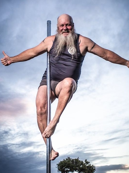 A grey bearded man in a singlet and little shorts balances on a pole