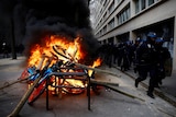 Black-clad riot police jog past a burning pile of what mostly appears to be electric bikes.