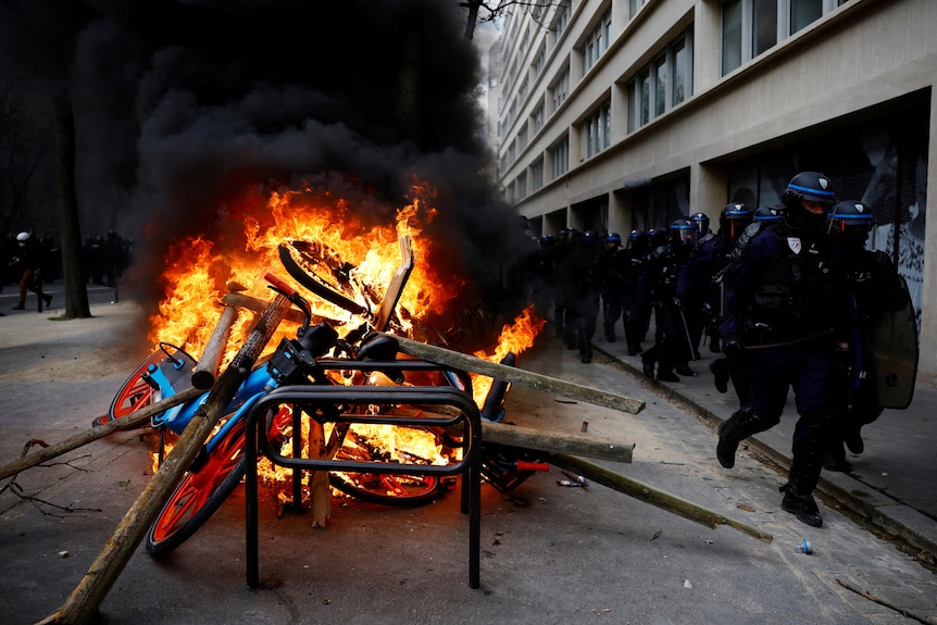 Black-clad riot police jog past a burning pile of what mostly appears to be electric bikes.