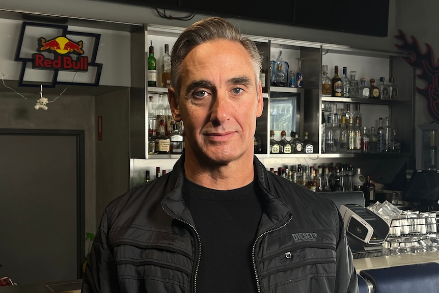 A man with silver hair stands in front of a bar stocked with alcohol