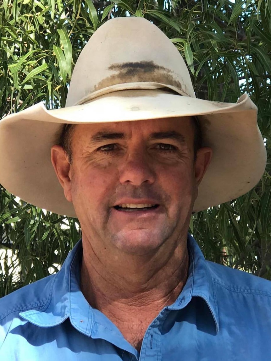 A man in a rural-style hat and blue shirt stands in front of a tree smiling.