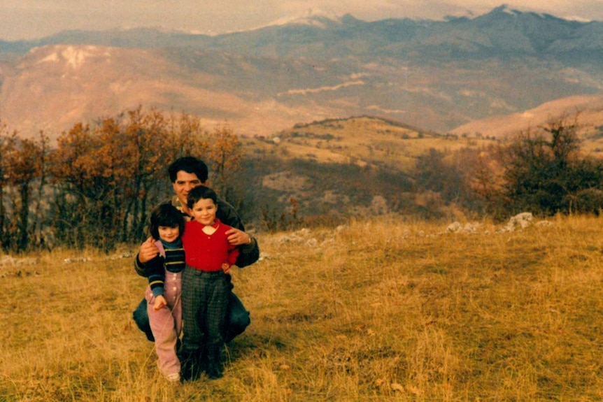 An old family photo of two girls with their father posing for a photo outside with mountains in the background.