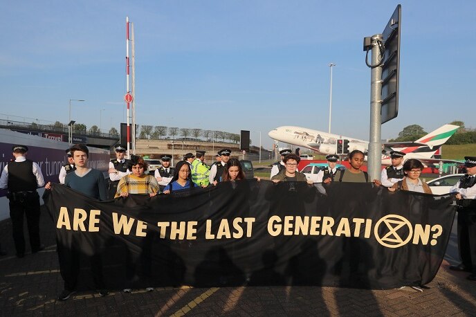 Group of teenagers hold a banner that says 'Are we the last generation' with a plane taking off in the background.