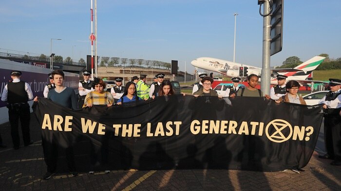 Group of teenagers hold a banner that says 'Are we the last generation' with a plane taking off in the background.