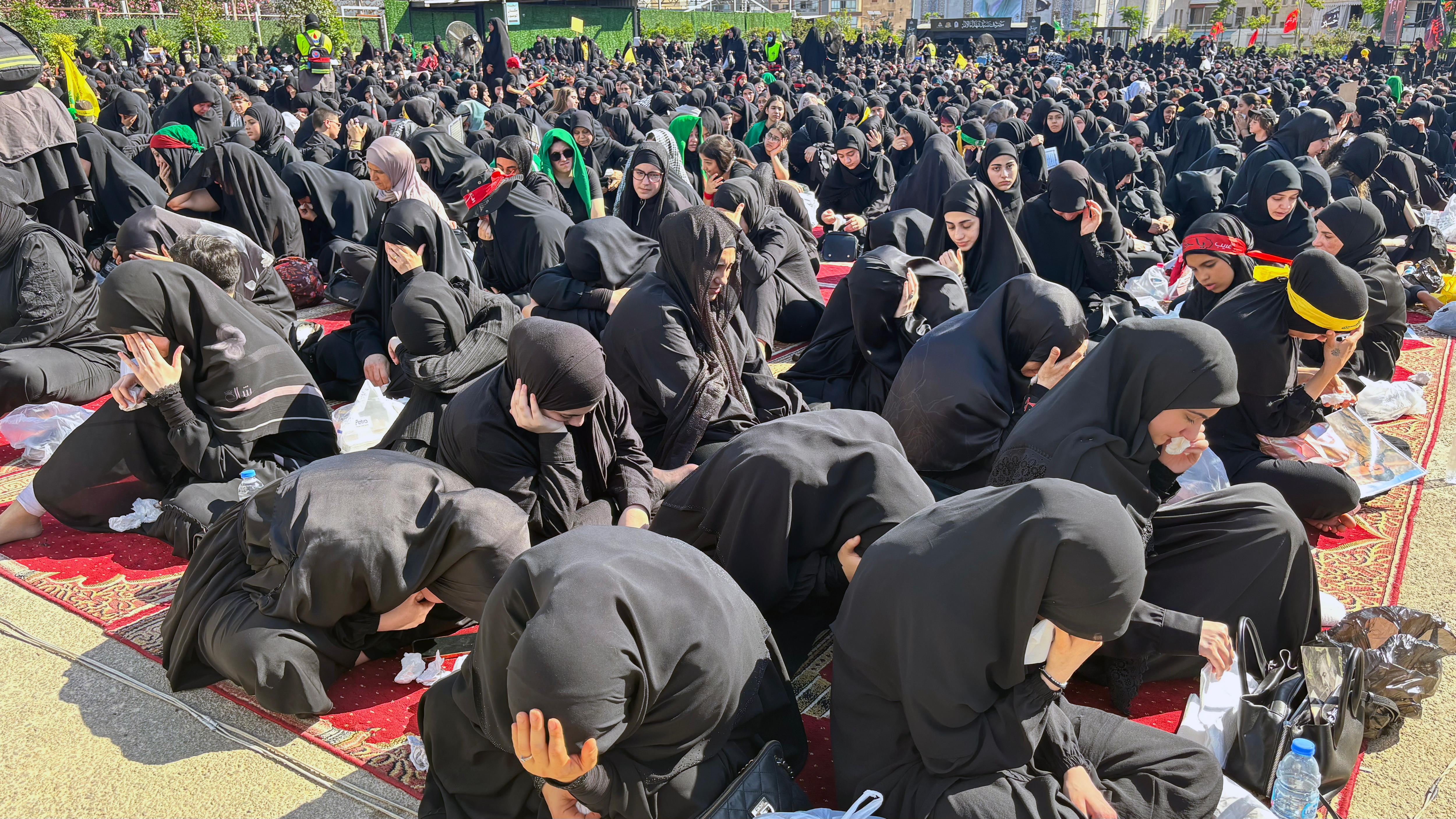 Dozens of women wearing black hijabs and robes are seen gathered and sitting on the ground, crying.