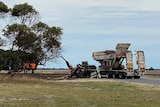 A smashed truck, missing it's cab, on the road close to a rail line. Two men stand in orange high-vis clothing near the truck.