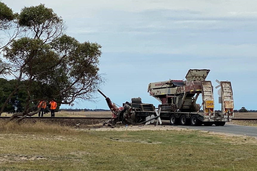 A smashed truck, missing it's cab, on the road close to a rail line. Two men stand in orange high-vis clothing near the truck.