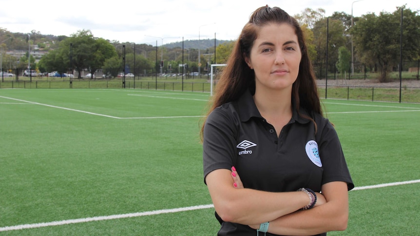 A female referee stands with her arms folded on the side of a football field.