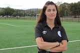 A female referee stands with her arms folded on the side of a football field.