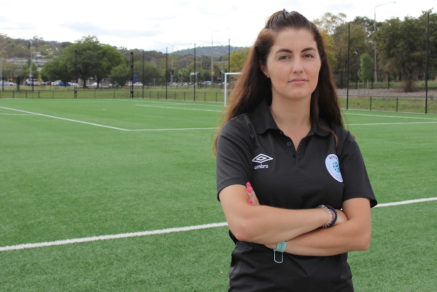 A female referee stands with her arms folded on the side of a football field.