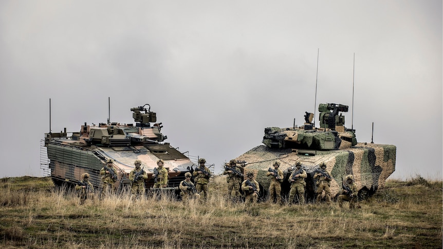A group of 12 soldiers in combat gear pose with their rifles in front of two large, heavy, tank-like combat vehicles in a field.