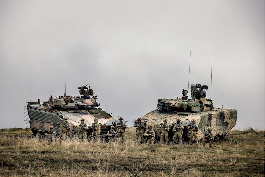A group of 12 soldiers in combat gear pose with their rifles in front of two large, heavy, tank-like combat vehicles in a field.