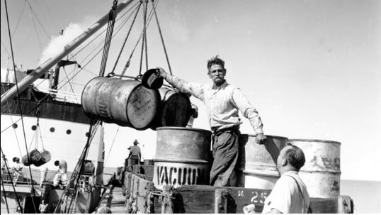 A black and white photo of a man with a large moustache loading drum barrels onto a large sail boat.