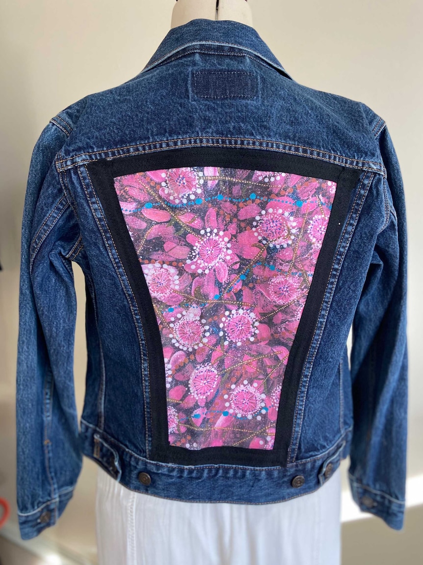 A denim jacket with a back panel featuring a pink and blue floral Indigenous art design