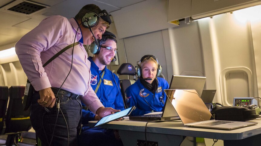 A man wearing a headset grins along with two people wearing NASA jumpsuits looking at laptop screens.