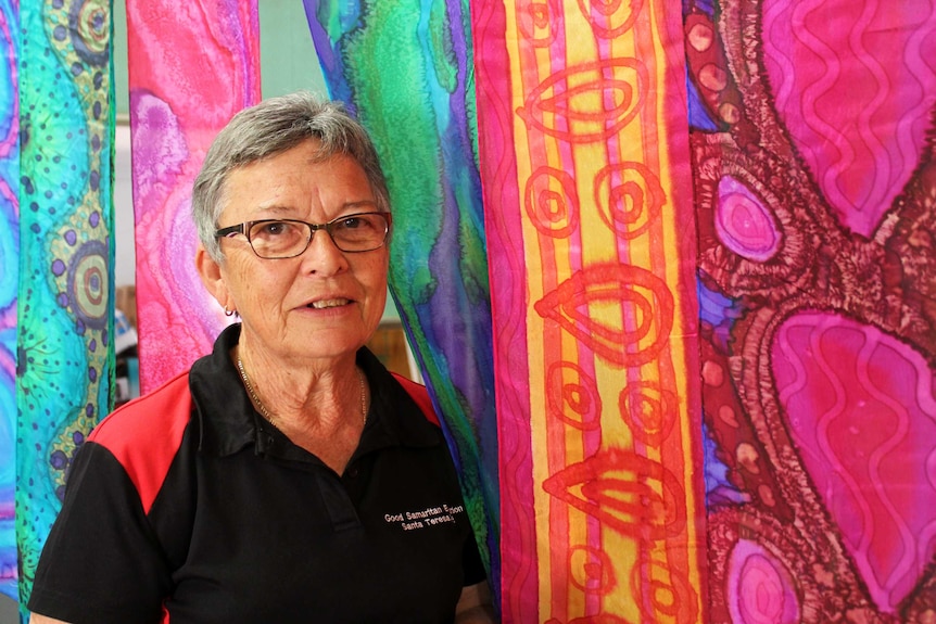 A woman with short grey hair and glasses stands in front of an Indigenous painting.