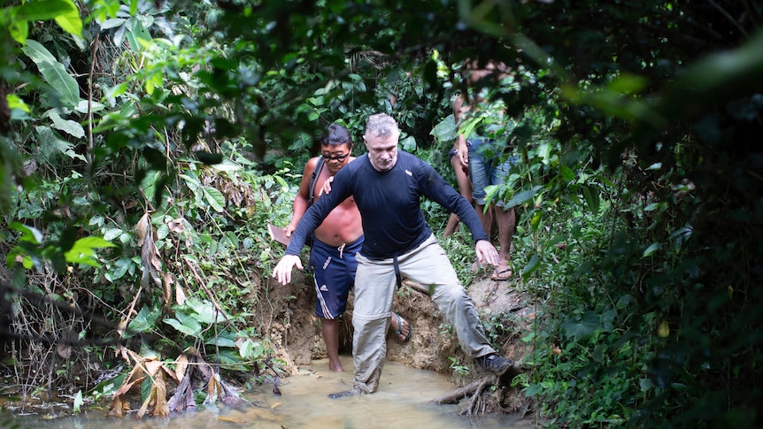Two men stepping into a narrow body of water, surrounded by lush green rainforest greenery.