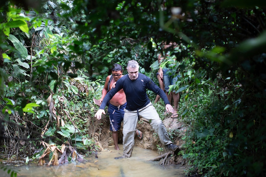 Two men stepping into a narrow body of water, surrounded by lush green rainforest greenery.